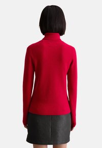Roter Rollkragenpullover mit geripptem Muster, taillierter Passform und langen Ärmeln, kombiniert mit einem strukturierten schwarzen Rock. Von hinten zeigt sich eine enge Passform.