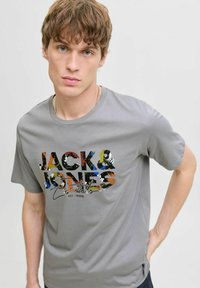 Harmaa puuvillapaita, jossa on monivärinen graafinen printti ja teksti "JACK & JONES Essentials" edessä. Lyhyet hihat ja pyöreä kaula-aukko.