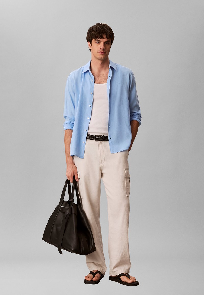 Jonge man in lichtblauw open overhemd over witte tanktop, beige cargobroek, zwarte gevlochten riem, zwarte teenslippers, houdend een grote zwarte leren tas.