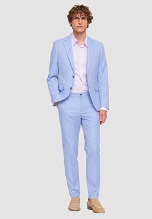Giovane uomo che indossa un completo azzurro chiaro con una camicia rosa pallido e mocassini beige, in piedi davanti a uno sfondo grigio chiaro uniforme.