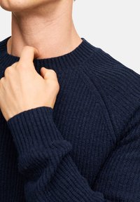 Marineblauer gerippter Strickpullover mit Rundhalsausschnitt, strukturiertem Stoff und langen Ärmeln mit schmalen Bündchen. Nahaufnahme einer Hand, die den Kragen anpasst.