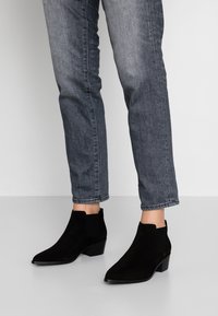 Schwarze Wildleder-Stiefeletten mit einer spitzen Zehenpartie und elastischen Seiteneinsätzen, mit einem klobigen Absatz, kombiniert mit grauen Jeans.