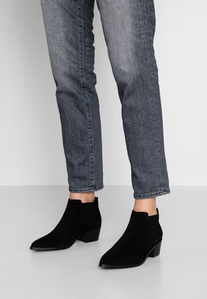 Stivali neri in suede alla caviglia con punta affilata e pannelli laterali elasticizzati, caratterizzati da un tacco spesso, abbinati a jeans grigi.