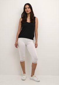 Zwart mouwloos topje met een v-hals, witte capri-leggings met kantafwerking, en witte sneakers. Toond tegen een effen achtergrond.