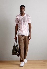 Camicia a righe a maniche corte di colore rosa chiaro e bianco, abbinata a pantaloni marroni. La modella tiene una borsa verde scuro e indossa sneakers bianche.