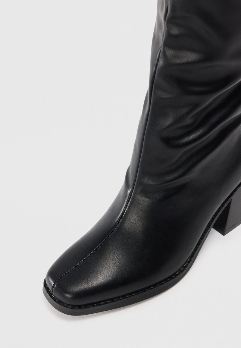 Bottine en cuir noir avec un bout carré, un design enroulé, des accents cousus et un talon bloc de hauteur intermédiaire. Texture lisse et finition brillante.