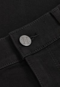 Close-up van zwarte denimstof met een metalen knoop gegraveerd met het woord "REISS" en gedetailleerd stiksel rond het knoopsgat.