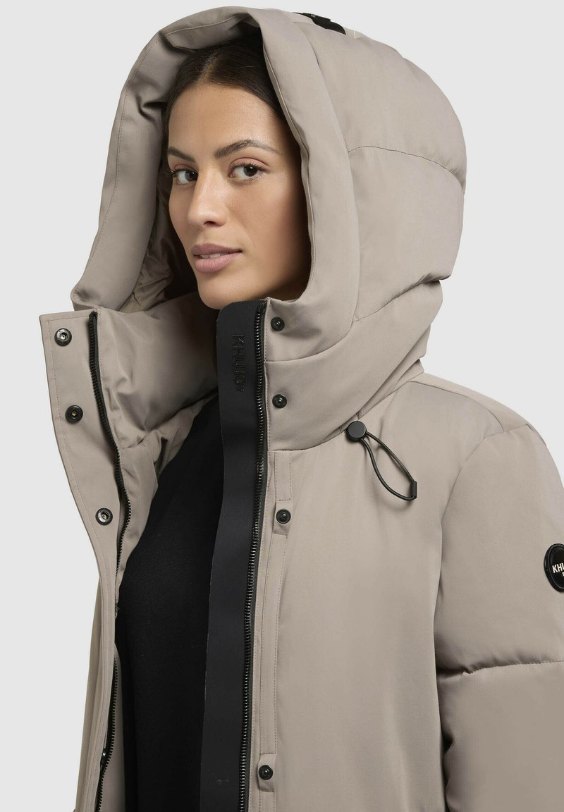 Beige Daunenjacke mit einer großen, plüschigen Kapuze. Merkmale einen schwarzen Reißverschluss, Druckknöpfe und eine verstellbare Schnur. Glatte Textur und minimalistisches Design.