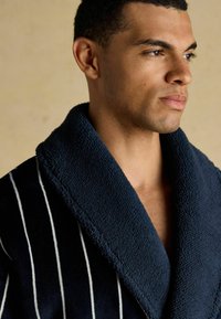 Man med kort hår klädd i en mörkblå badrock med vita ränder och en tjock texturerad sjalkrage, som ser åt sidan mot en enkel bakgrund.