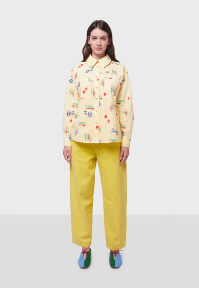 Camisa amarilla de botones con patrones abstractos coloridos, combinada con pantalones amarillos de pierna ancha. Los zapatos son azules y verdes con cordones rojos.