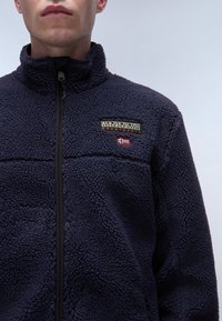 Marineblaue Fleecejacke mit hohem Kragen, Reißverschluss und strukturierter Oberfläche. Gesticktes Logo und Flaggen-Details auf der Brust.