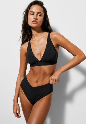 Kobieta o długich ciemnych włosach, ubrana w czarny strój kąpielowy z dekoltem w serek i wysokim stanem, pozująca z jedną ręką na biodrze na jednolitym tle.