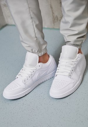 Personne portant des baskets blanches Nike avec lacets, debout sur un sol bleu clair tacheté, vêtue d'un pantalon gris clair avec des poignets élastiques.