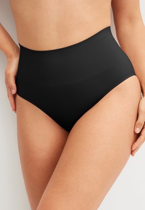 Vrouw draagt hoge taille zwarte naadloze corrigerende slips, handen rustend bij de heupen, neutrale achtergrond.