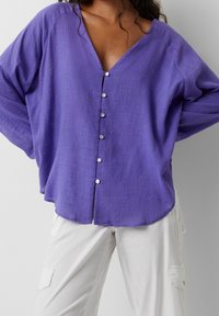 Blusa de lino morada con un escote en V profundo, corte holgado y cinco botones frontales. Presenta mangas anchas y dobladillo curvado. Combinada con pantalones claros.