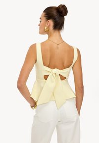 Haut peplum en jaune clair, présentant un grand nœud dans le dos, des découpes profondes et des bretelles larges, associé à un pantalon blanc.