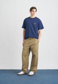 Mornarsko modra majica s kratkimi rokavi z majhnim rumenim logotipom na prsih, v kombinaciji s svetlimi širokimi hlačami in belimi supergami.