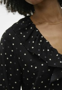 Schwarze Bluse mit weißen und helllila Punkte. Sie verfügt über einen gefransten Kragen, strukturierten Stoff und ein V-Ausschnitt-Design.