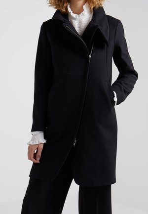 Vrouw draagt een zwarte jas met rits over een witte blouse met ruches aan kraag en manchetten, staand met één hand in de zak.