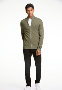 Khaki zip-up tröja i texturerat tyg, med hög krage och ribbade muddars, i kombination med svarta byxor och vita sneakers.