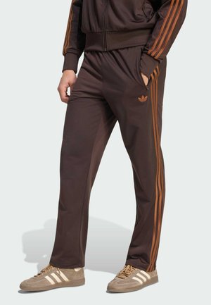 Persona che indossa pantaloni della tuta marroni Adidas con strisce arancioni, giacca abbinata e scarpe sportive Adidas beige, in piedi con una mano in tasca.