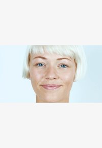 Närbild av en person med kort, rakt platina-blont hår, blå ögon och en slät hy, som ler mot en ljus bakgrund.