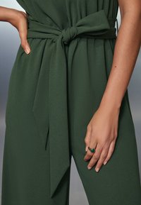 Grön overall med ett knytbart midjeband, tillverkad av mjukt, texturerat tyg. En hand dekorerad med en guldring med grön sten vilar vid sidan.