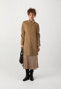 Brūna rāvējslēdzēja jaka no teksturēta auduma ar apkakli, divām priekšpusē kabatām un garām piedurknēm. Valkāta pār bēšu svārkiem un melniem zābakiem līdz ceļiem.