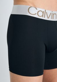 Fekete pamut boxer alsónadrág, ezüst derékrésszel, arany betűkkel a Calvin felirattal. Sima tapintás és testhez simuló fazon.