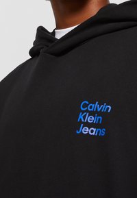 Sudadera negra con capucha hecha de algodón con una textura suave. Presenta un logotipo de texto azul "Calvin Klein Jeans" en el frente. Diseño clásico con capucha.