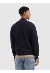 Marineblauwe gebreide trui met een geribbeld ontwerp, kraag en manchetten. Draagt over een lichtbruine shirt, gecombineerd met lichtblauwe versleten jeans.