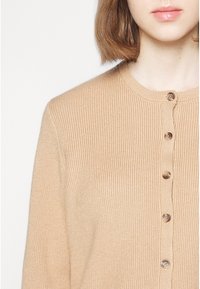 Femme portant un cardigan en maille côtelée beige avec cinq boutons marron, visible du cou au milieu du torse sur un fond blanc uni.