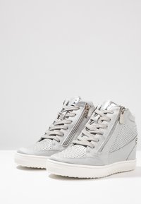 Grå kile-sneakers med perforert semsket skinn, metalliske detaljer, snøring foran og glidelåser på siden for enkel påkledning. Flate hvite gummisåler.