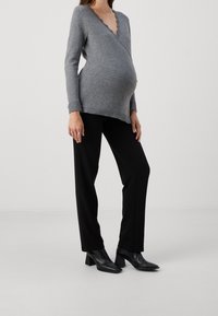 Kobieta w ciąży, ubrana w szary długi rękaw, czarne spodnie i czarne botki na obcasie, stoi na jednolitym jasnym tle.