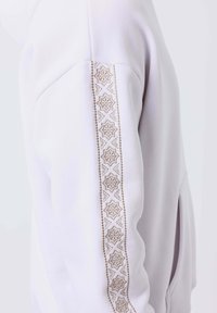 Vêtement blanc à manches longues avec un motif vertical d'étoile géométrique brodée beige le long du bras extérieur.
