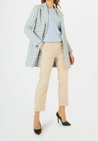 Manteau à carreaux en bleu clair et blanc avec des manches longues sur un haut bleu clair, porté avec un pantalon beige et des talons hauts noirs.