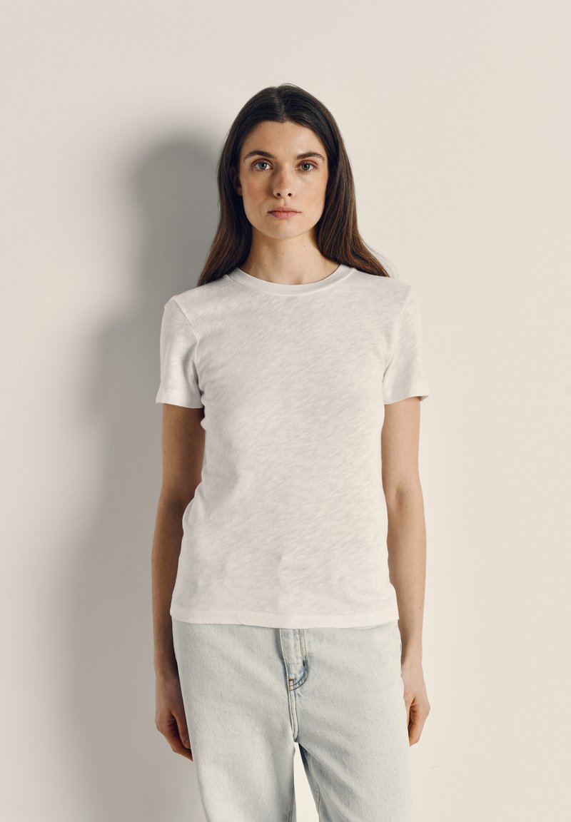 Mujer con cabello largo y castaño, vistiendo una camiseta blanca lisa y jeans azul claro, de pie contra una pared beige, mirando hacia adelante.