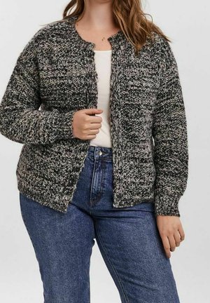 Femme portant un cardigan texturé noir et blanc par-dessus un haut blanc, associé à un jean taille haute bleu, debout devant un fond uni.