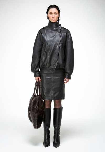 Žena stojí v čiernej koženej bunde, zladená s koženou sukňou, vysokými čižmami po kolená a drží veľkú tmavú kožušinovú kabelku.