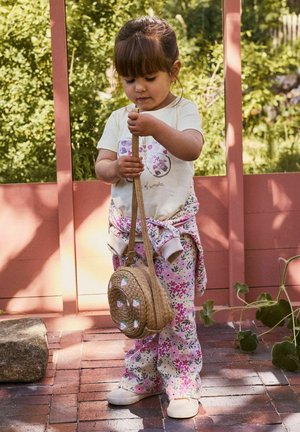 Jeune fille en pantalon à fleurs et chemise blanche tenant un sac rond tressé, debout sur un patio en briques avec de la verdure en arrière-plan.