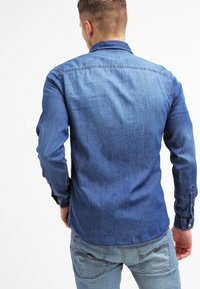 Camicia in denim con bottoni, toni di blu in gradiente, maniche lunghe, vestibilità su misura, colletto piccolo e dettagli di cucitura visibili sulle spalle.