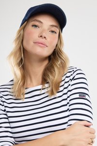 Blond kvinde med bølget hår iført en marineblå kasket og en hvid skjorte med sorte vandrette striber, arme krydset, neutral udtryk.