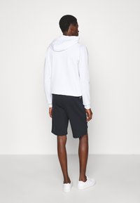 Felpa bianca da uomo con cappuccio regolabile, abbinata a pantaloni corti neri. La felpa ha polsini a coste; il modello indossa sneakers bianche. Design minimalista.