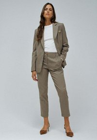 Blazer de cuadros marrón con pantalones a juego, blusa blanca debajo y zapatos de tacón marrones. Incluye un pin en la solapa y bolsillos laterales.