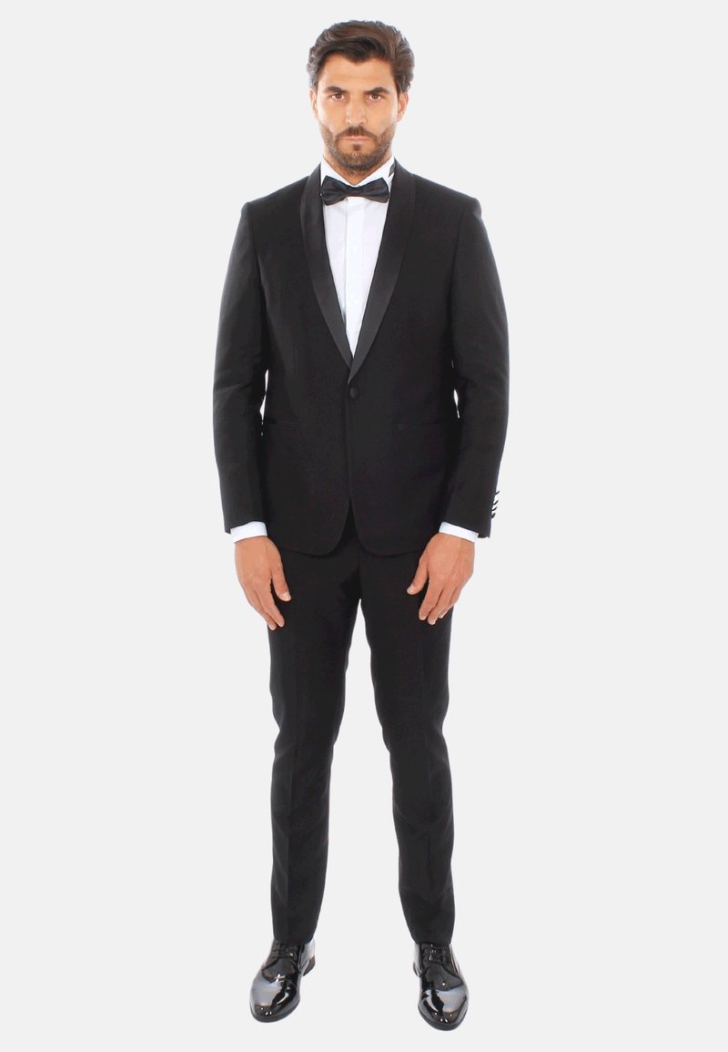 Tuxedo nero con rever in satin, design aderente, camicia bianca e papillon nero. Scarpe eleganti nere lucide. Posizione di fronte a uno sfondo bianco.
