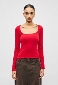 Dugih rukava crveni top sa kvadratnim izrezom, ukrašen malom mašnom u sredini. Mekani, pripijeni materijal glatke teksture.