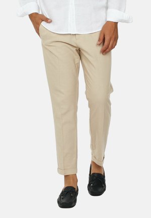 Uomo che indossa pantaloni aderenti beige con orli risvoltati, camicia bianca e mocassini neri, che cammina con una mano in tasca.