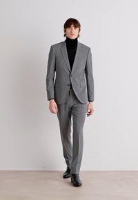 Abito grigio con vestibilità su misura, caratterizzato da un chiusura a un bottone, abbinato a un dolcevita nero e scarpe nere. Tessuto liscio.