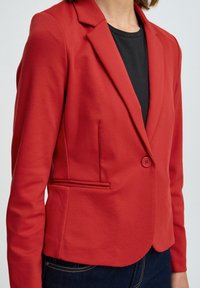 Blazer rosso in tessuto liscio, con colletto a rever, chiusura a un bottone e tasche laterali, abbinato a un top nero.