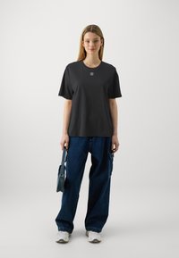 Black short-sleeve t-shirt with a round neck, featuring a subtle logo on the chest, paired with oversized blue jeans and white sneakers.